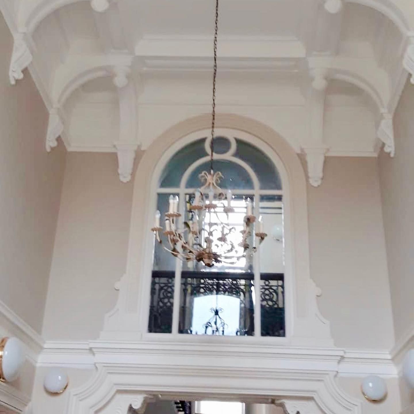 High-ceilinged room with large arched window and chandelier. Ornate white molding on beige walls.
