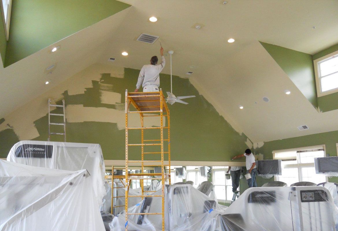 Two painters painting the interior of a gym with a high ceiling. Equipment is covered in plastic.