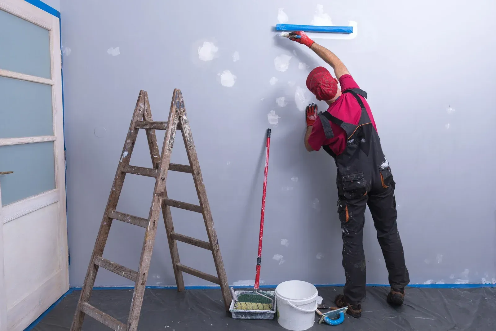 Person in overalls painting a wall with a roller, next to a ladder and paint supplies.