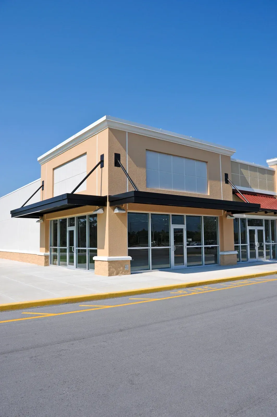 Empty beige retail building with glass windows and black awnings against a clear blue sky.