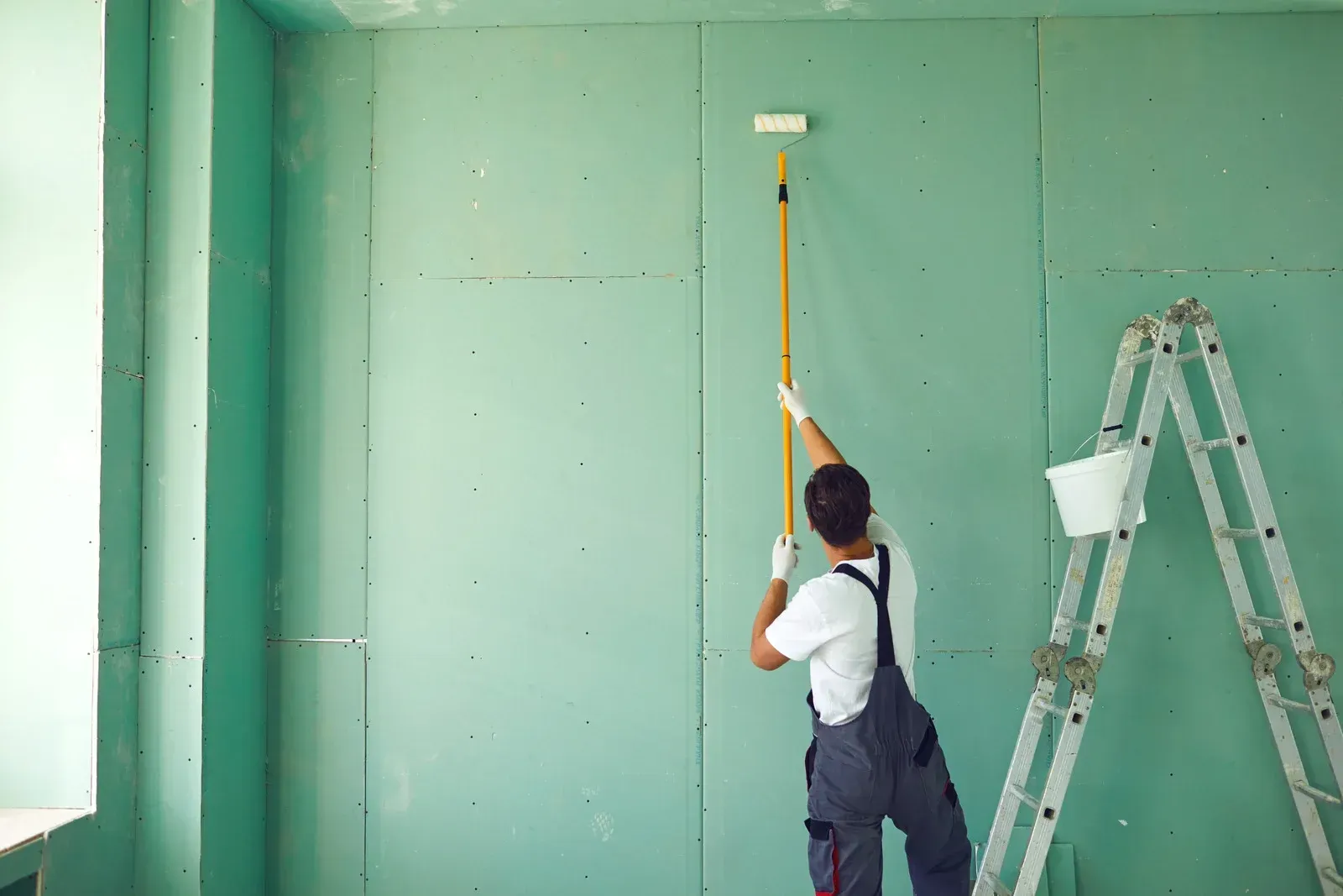 Person paints a green wall with a roller on a long handle, standing near a ladder in a room.