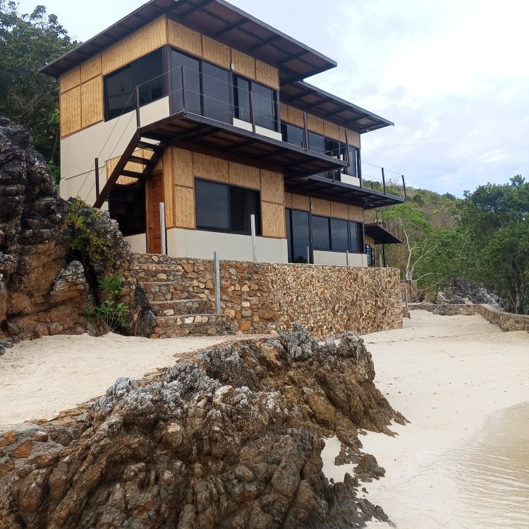 A large house sits on top of a rocky hill next to a beach