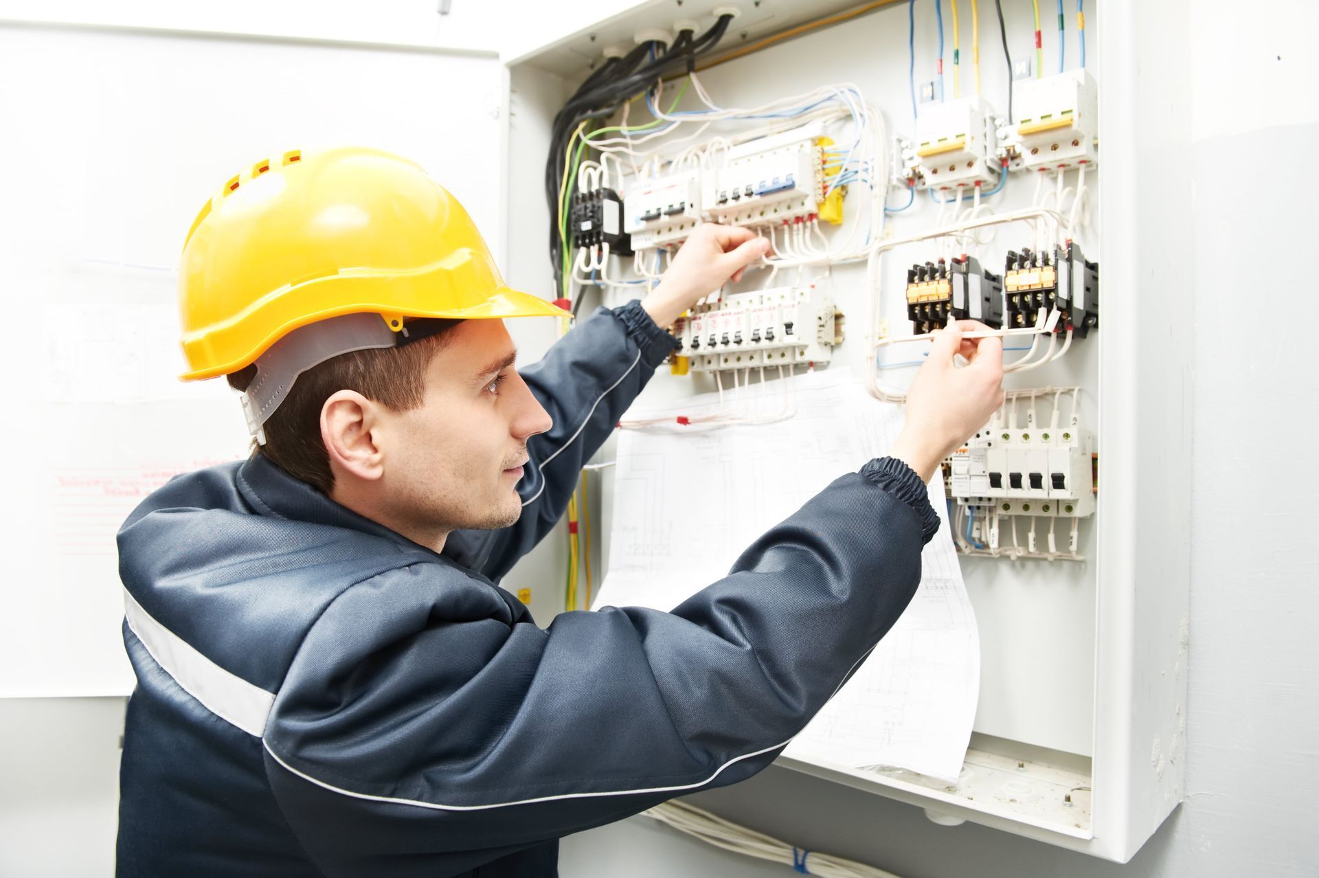 Electrician in a yellow hard hat inspecting wiring inside an open electrical control panel.