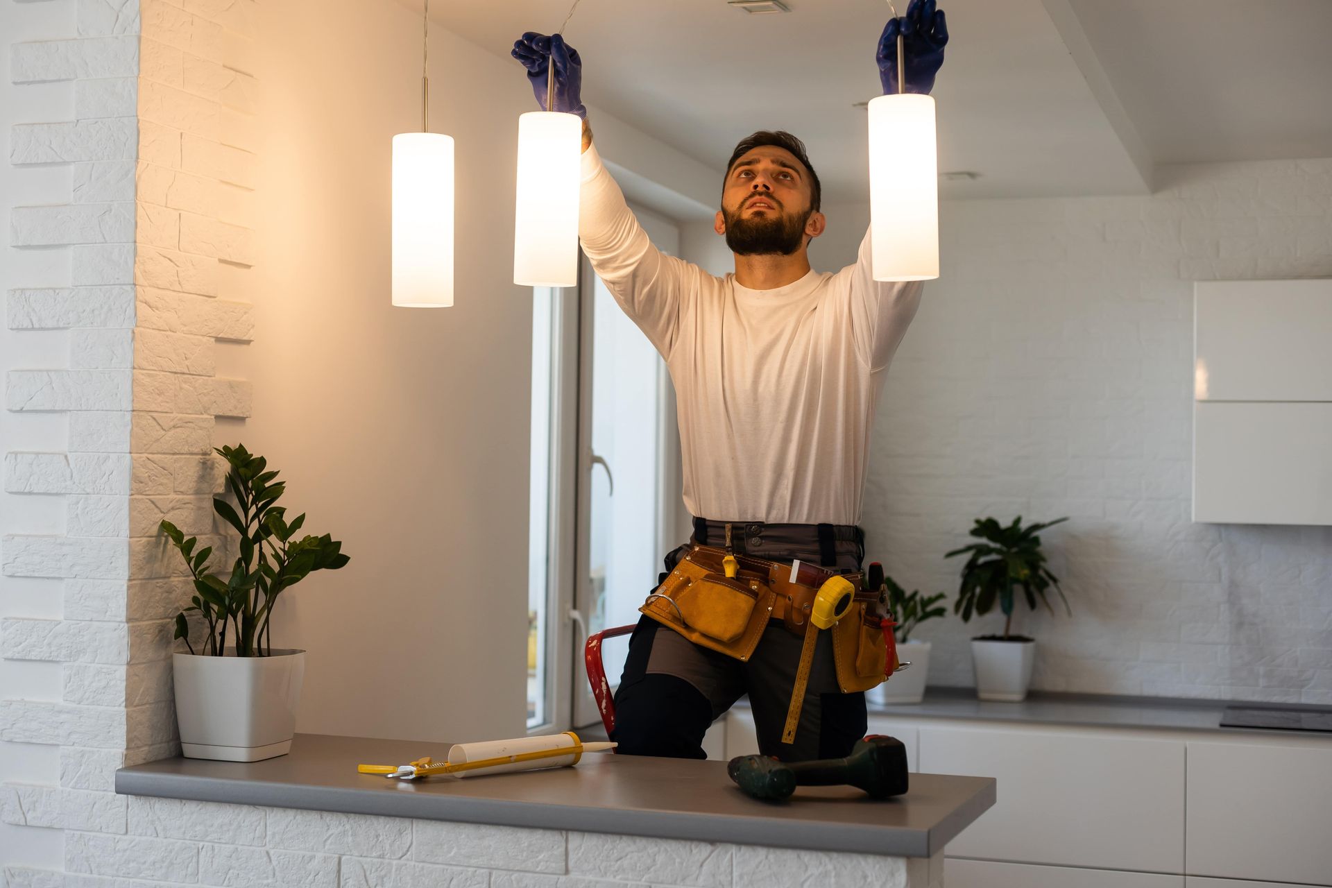 Electrician installing three pendant lights in a modern kitchen, standing on a counter with tools nearby