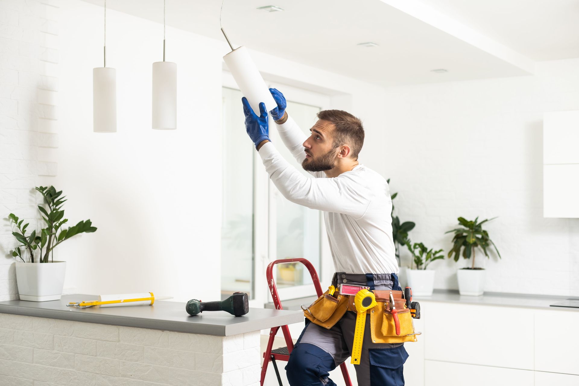 Electrician installing a ceiling light in a bright kitchen, using a drill and tool belt.