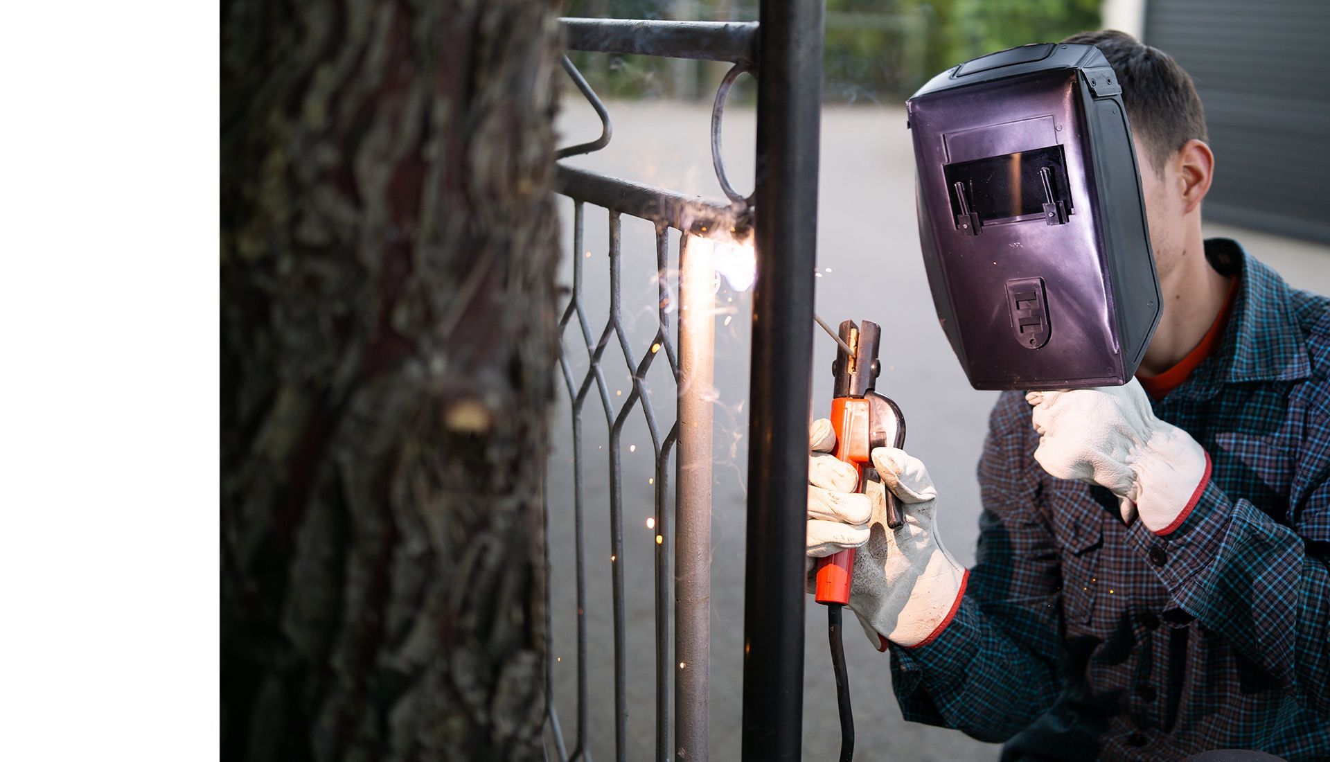 A man wearing a welding mask is welding a metal fence.