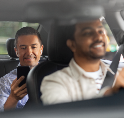 Man in the back seat using phone; driver smiling behind the wheel, inside a car.