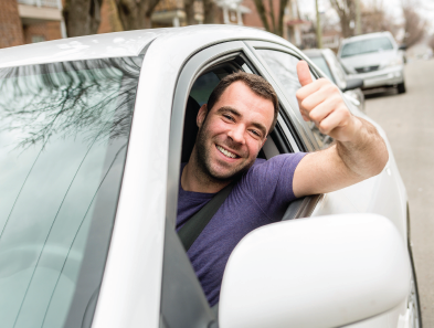 Man in car giving a thumbs-up, smiling. Wearing seatbelt. Silver car, street in background.