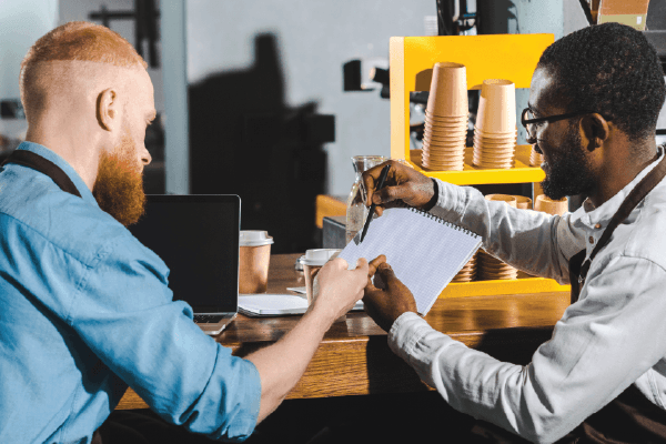 Two cafe workers reviewing papers together, one red-haired, one Black, wearing aprons.