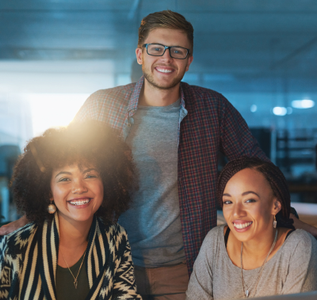 Three smiling coworkers in an office: two women in front, one man behind, all looking at the camera.