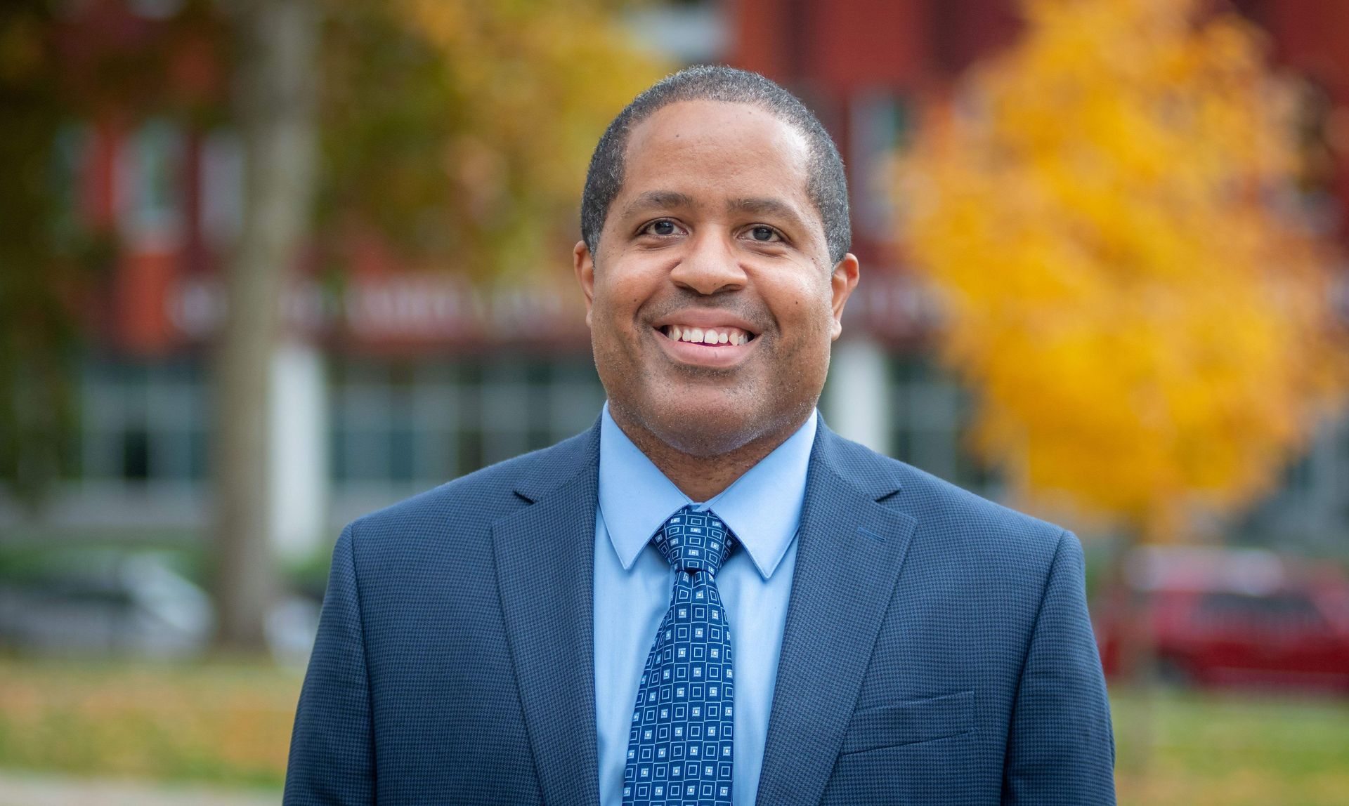 Man in a blue suit smiles outdoors, a building and fall foliage in the background.