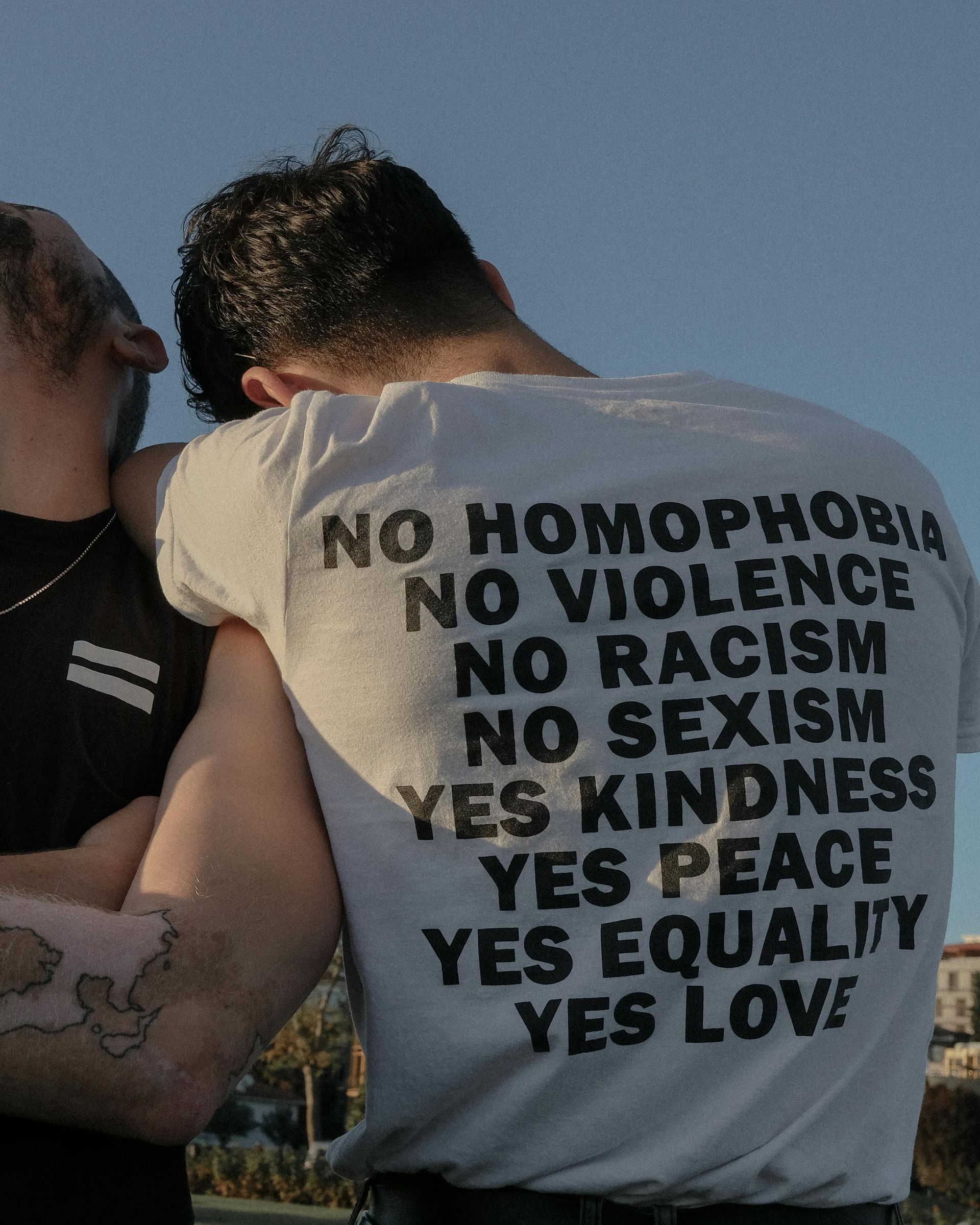 Person wearing a t-shirt with text promoting kindness, peace, equality, and love. Another person is embracing them.