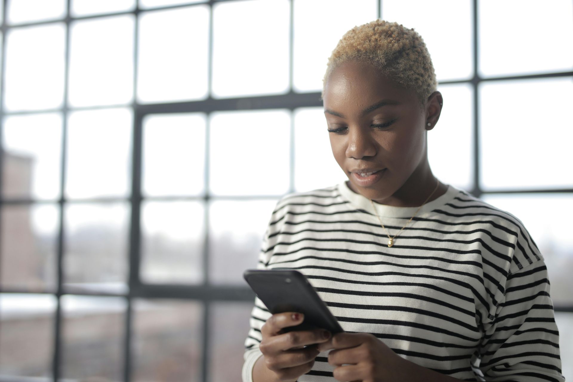 Woman with short blonde hair looking at a smartphone, standing near a large window.