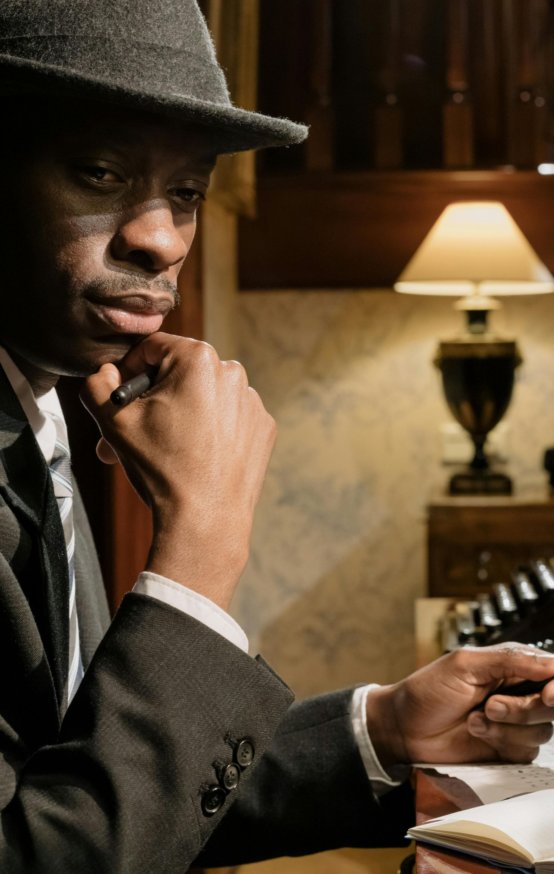 Man in fedora, writing at desk with lamp, thoughtful expression.