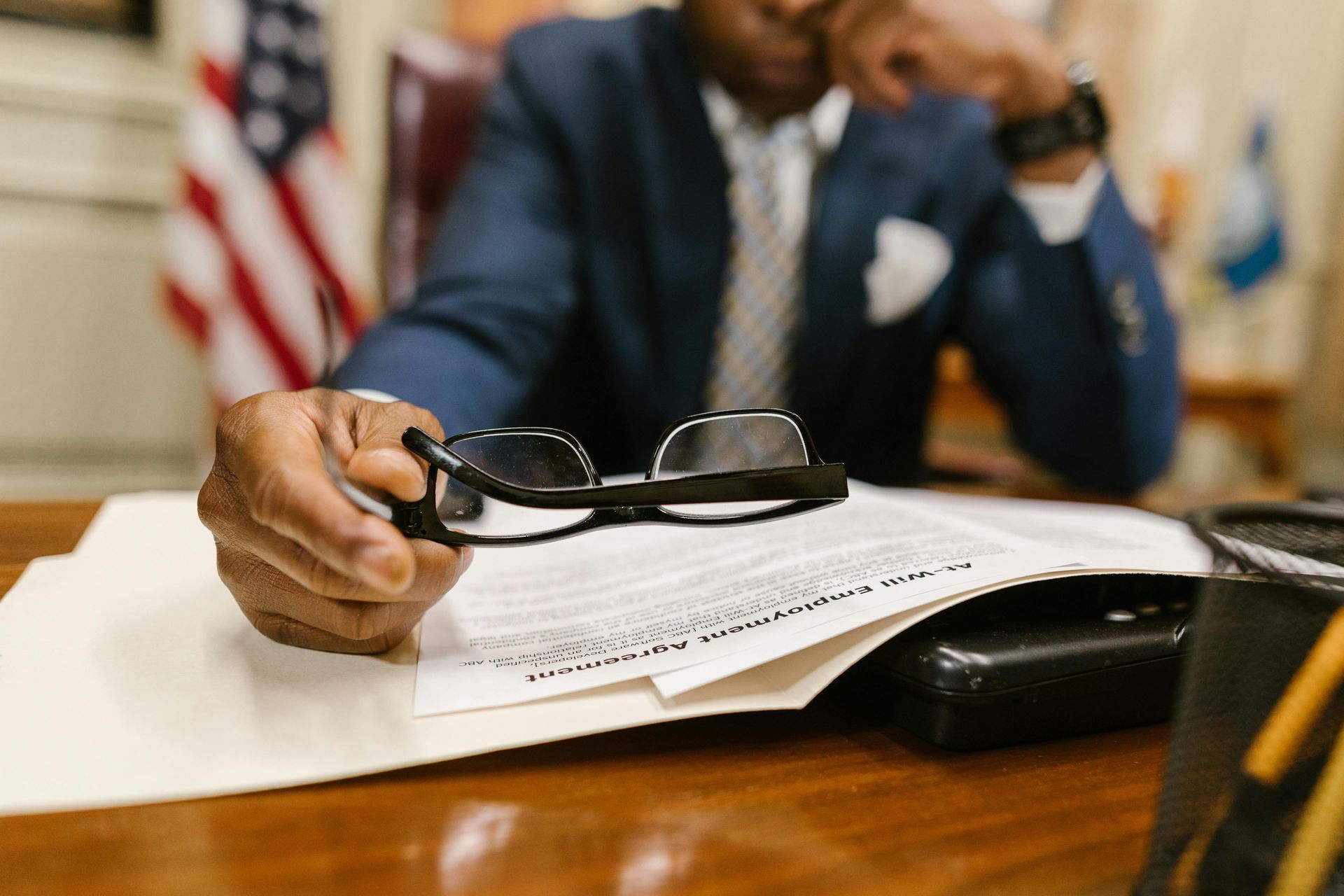 Person in a suit holding eyeglasses, looking at documents on a desk with a US flag in the background.