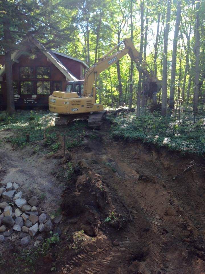 A yellow excavator is digging a hole in the woods in front of a house.