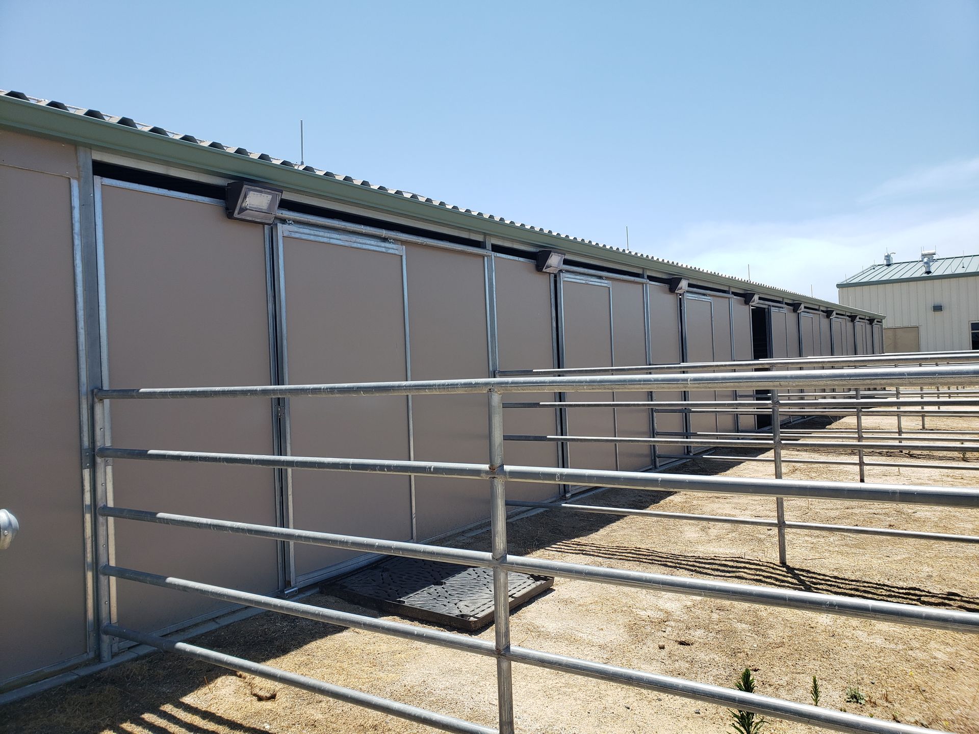 Tan horse stalls with silver metal doors, a steel rail fence, and a light blue sky.