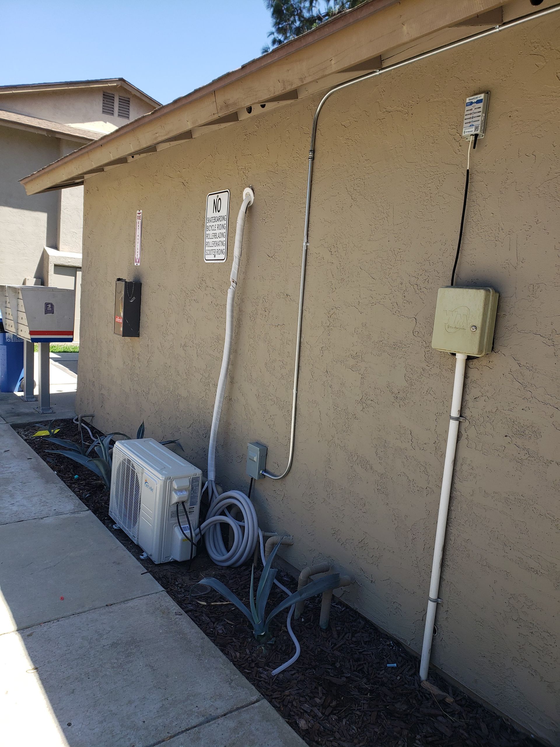 Exterior wall with electrical boxes, wires, and air conditioning unit near a sidewalk.