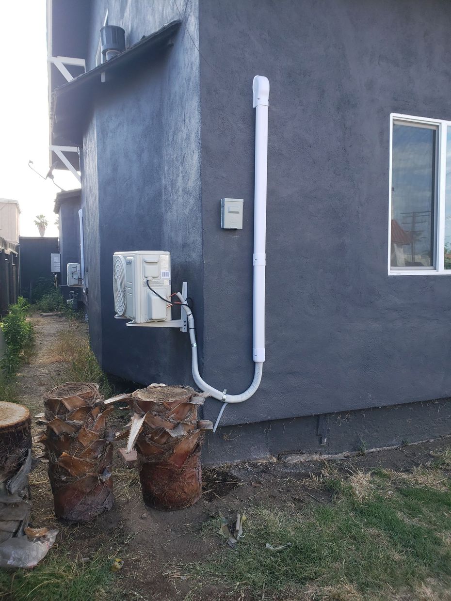 Air conditioner unit on a dark gray stucco wall with white piping. Side yard with tree stumps.