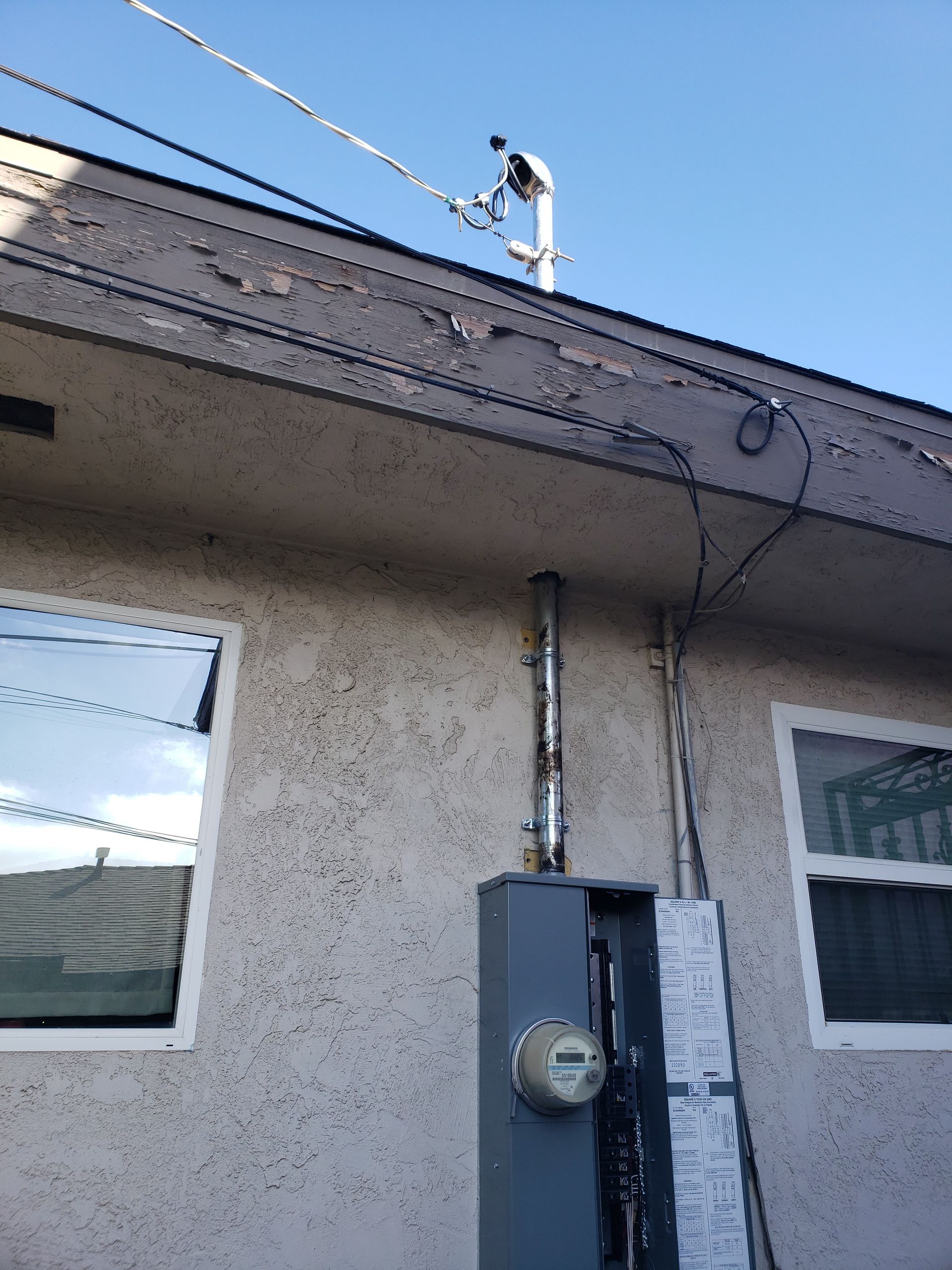 Exterior wall with electrical meter box, conduits, and antenna against a blue sky.