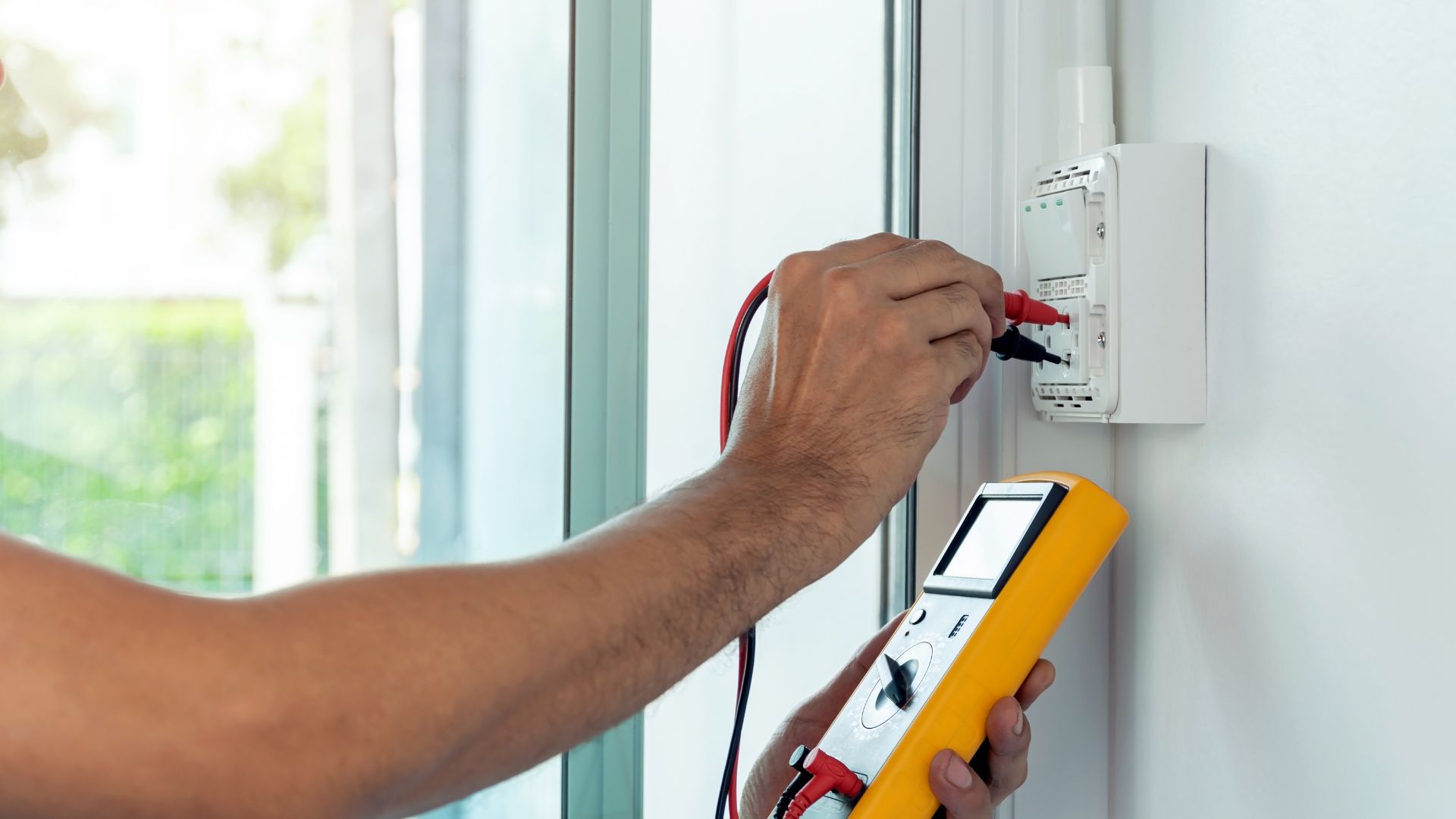 Person using a yellow multimeter to check electrical outlet's voltage on a white wall.