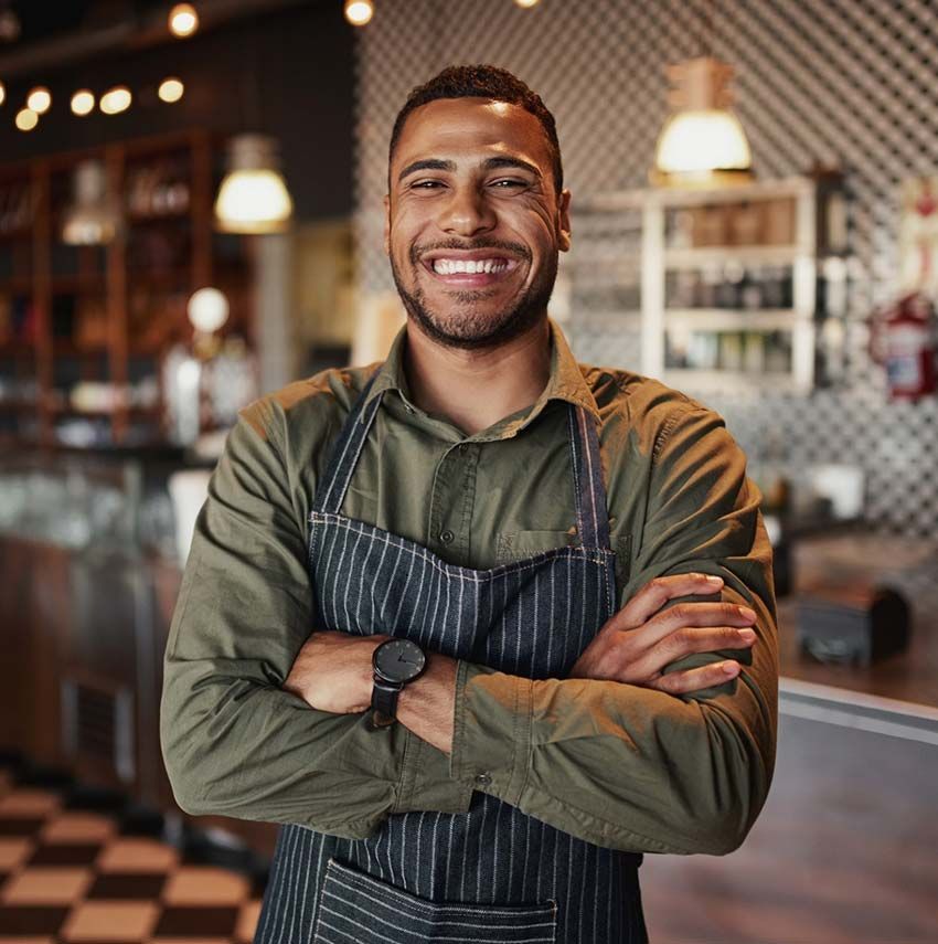 A man in an apron is standing in a restaurant with his arms crossed and smiling.