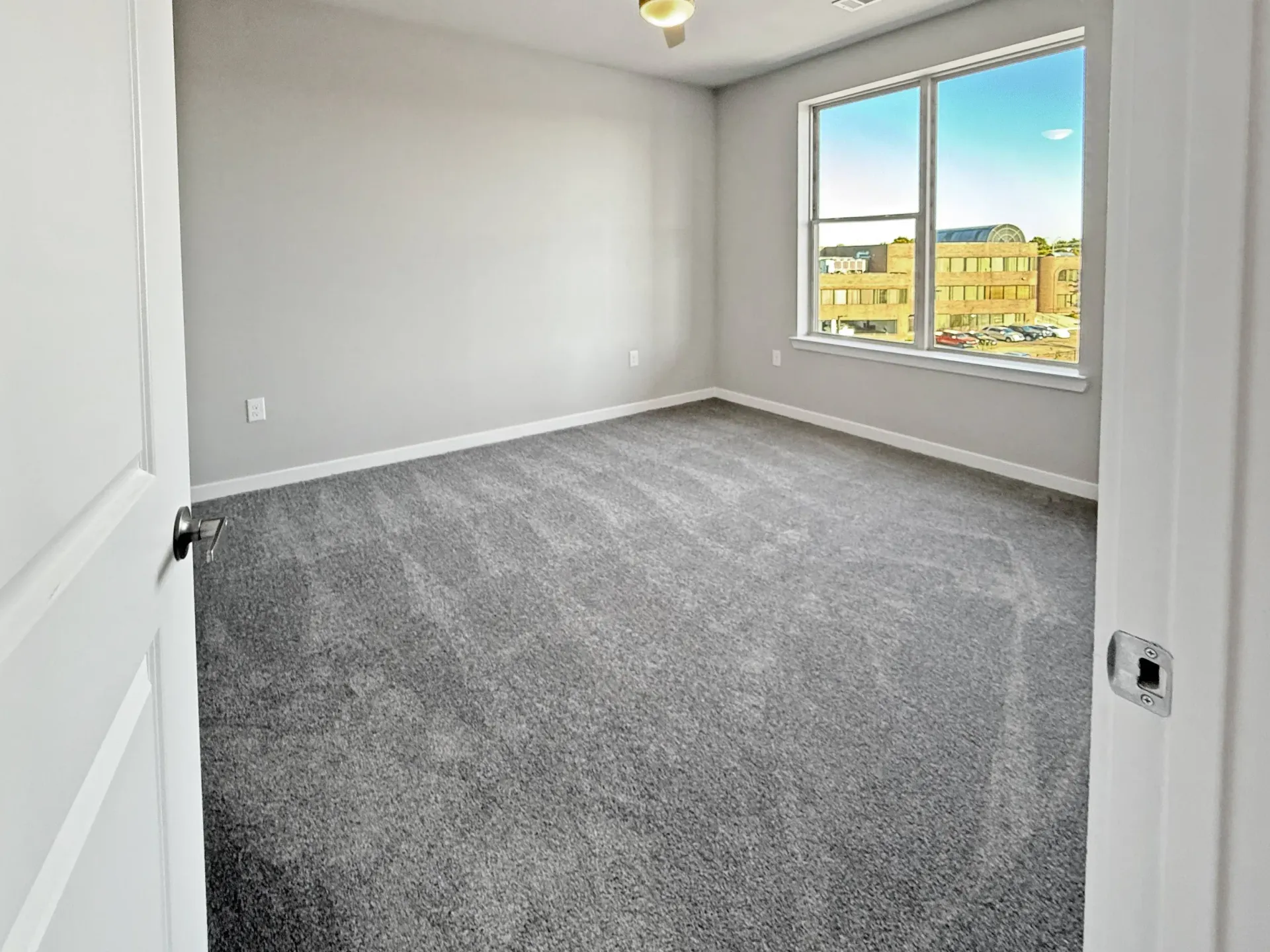 Empty bedroom with gray carpet and light gray walls, large window with city view.