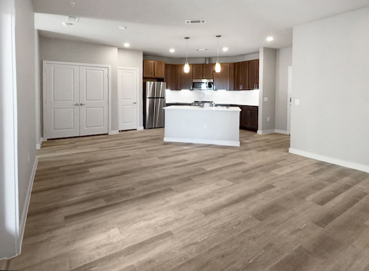 Open-concept living space with kitchen and wood-look flooring at Urban Village at Rosehill, featuring brand-new apartments in Garland, TX. Neutral walls, dark cabinets, and a ceiling fan are visible.