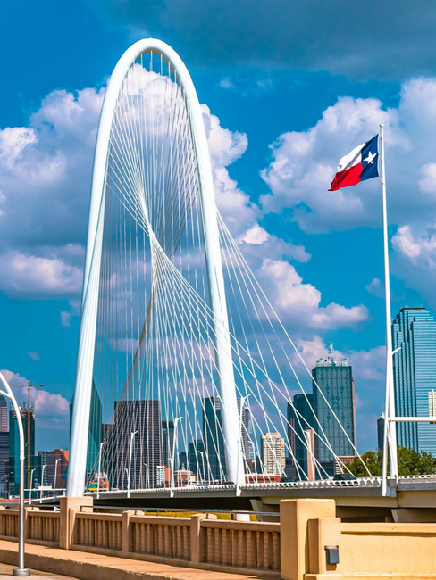 Dallas skyline with white Margaret Hunt Hill Bridge under a blue sky, Texas flag waving.