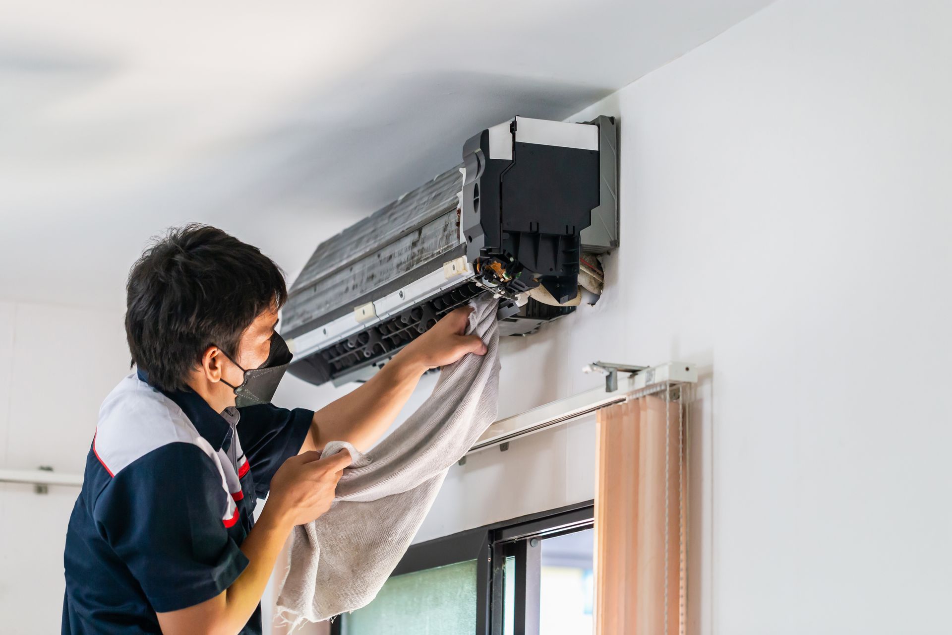 A man wearing a mask is cleaning an air conditioner with a towel.