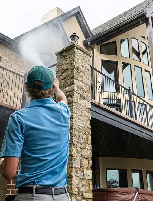 Person in blue shirt power washing a stone column on a house with large windows.