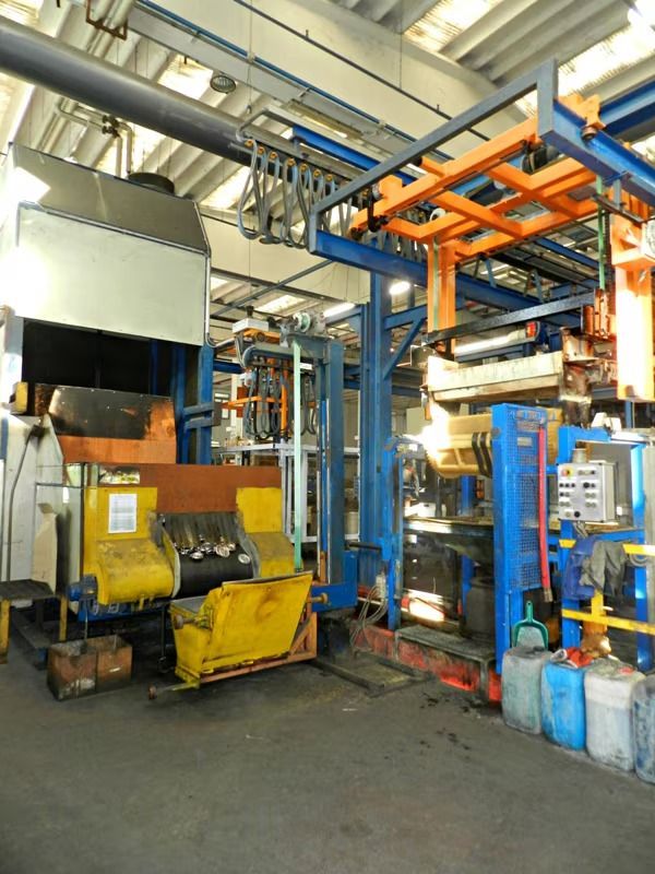 Interno di una fabbrica industriale con macchinari, tubi e container blu e arancioni su un pavimento di cemento.