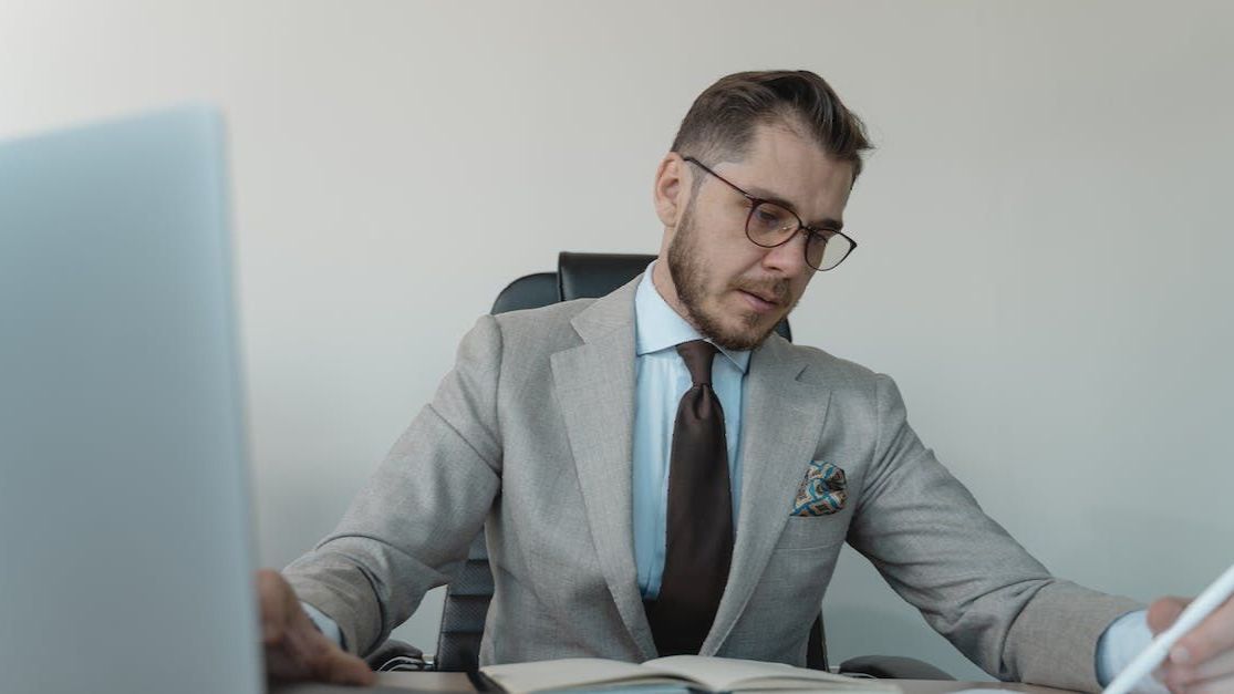 A man in a suit and tie is sitting at a desk in front of a laptop computer.