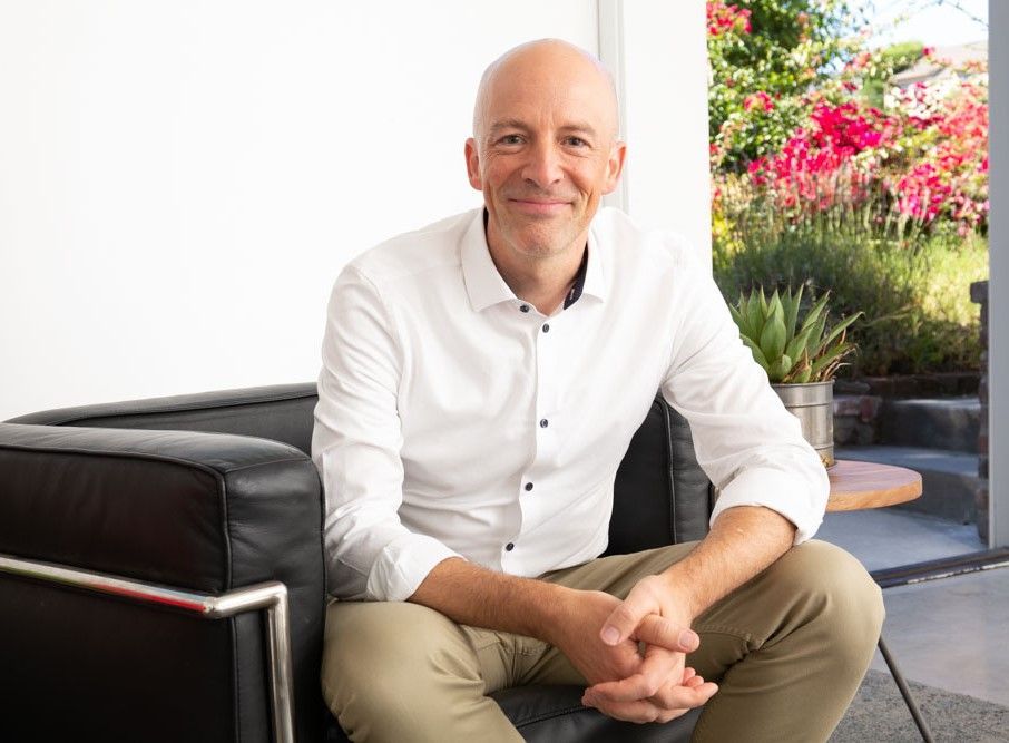 Man with shaved head smiles, seated in a black chair, wearing a white shirt and khaki pants