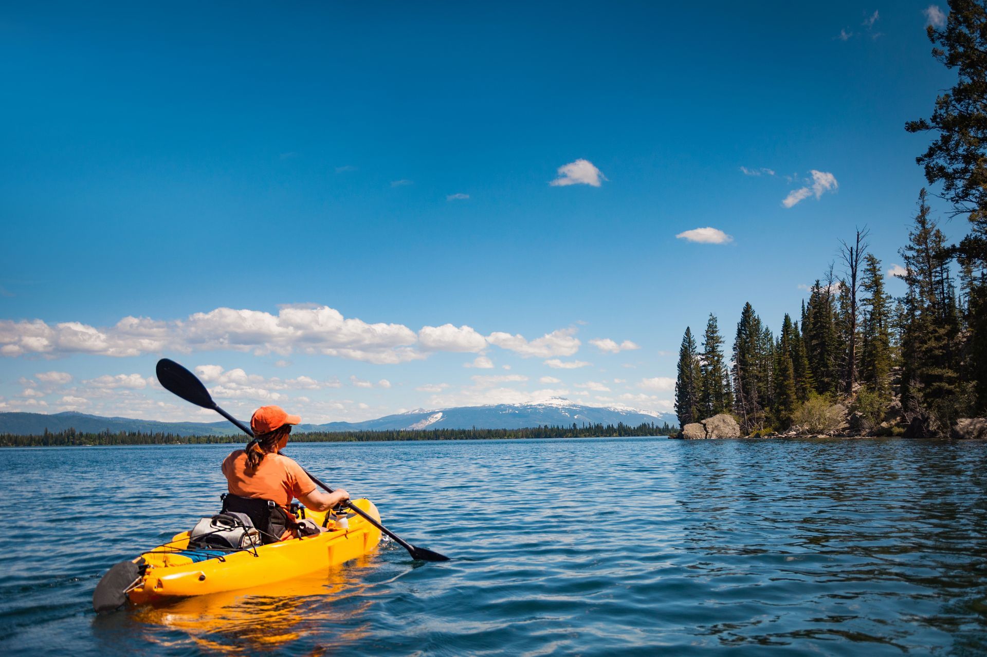 Person kayaking in a yellow kayak on blue water; bright blue sky with trees in background.