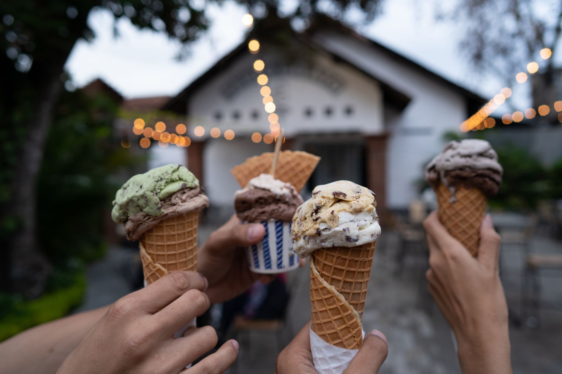 Hands holding ice cream cones in front of a white building with string lights.