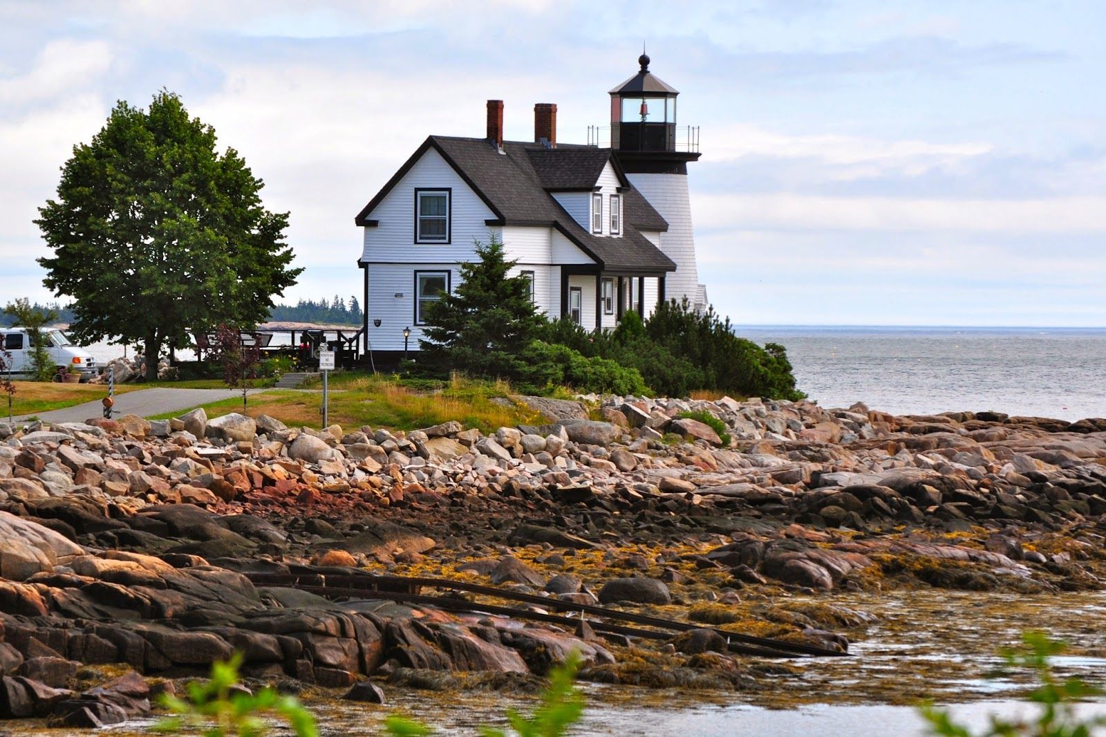 Lighthouse on a rocky coast, with a white building and black tower. The scene is near the ocean with a few trees.