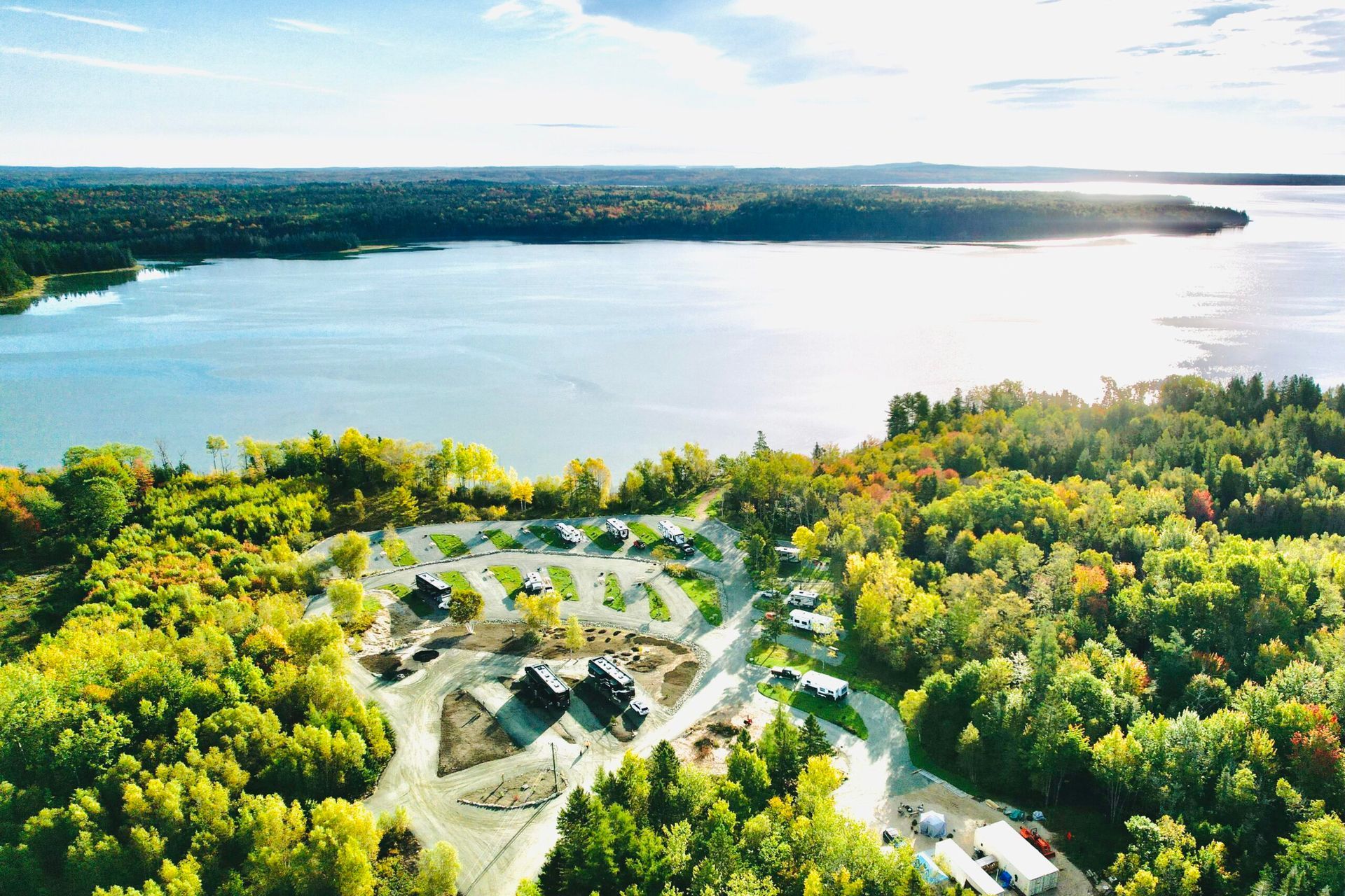 Aerial view of a campground next to a lake surrounded by trees in autumn foliage. The sun shines on the water.