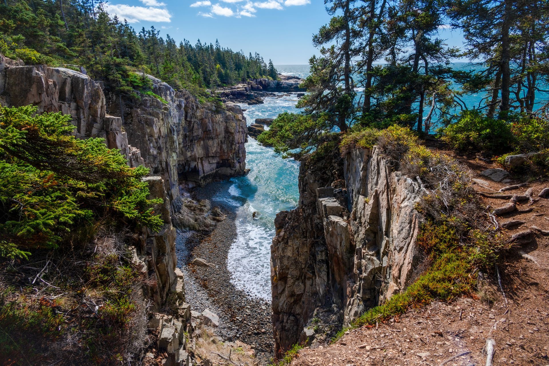 Rocky coastline with a narrow inlet of water between cliffs, trees on the cliff edges, and a bright blue sky.