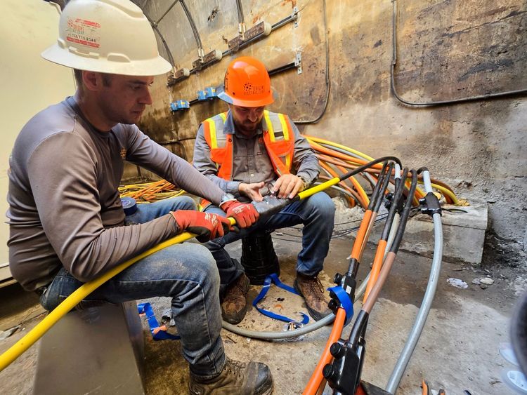 Two workers wearing hard hats and safety gear work with thick electrical cables in an underground utility space.
