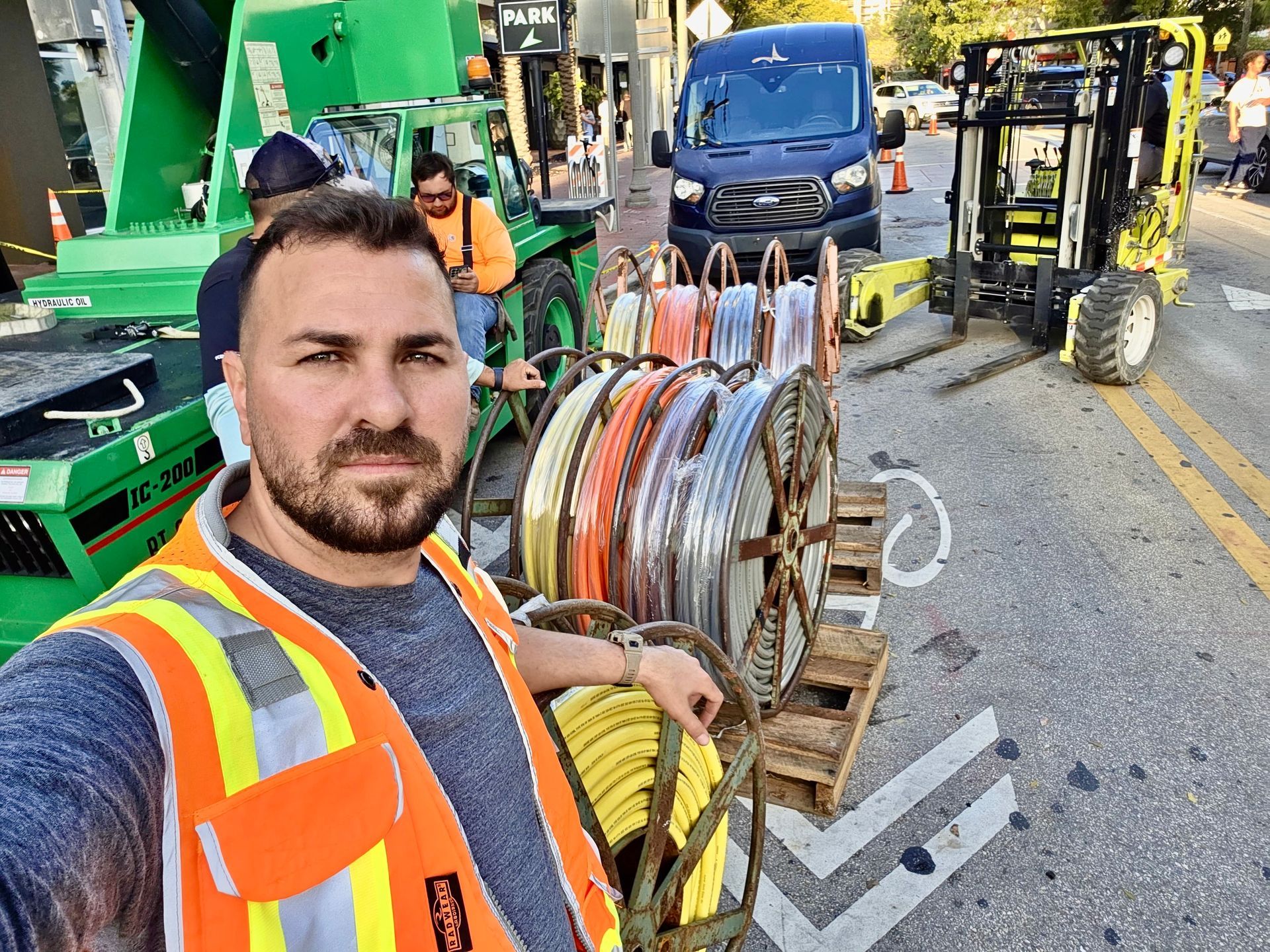 A construction worker wearing a high-visibility vest takes a selfie on a street with cable spools and heavy machinery.