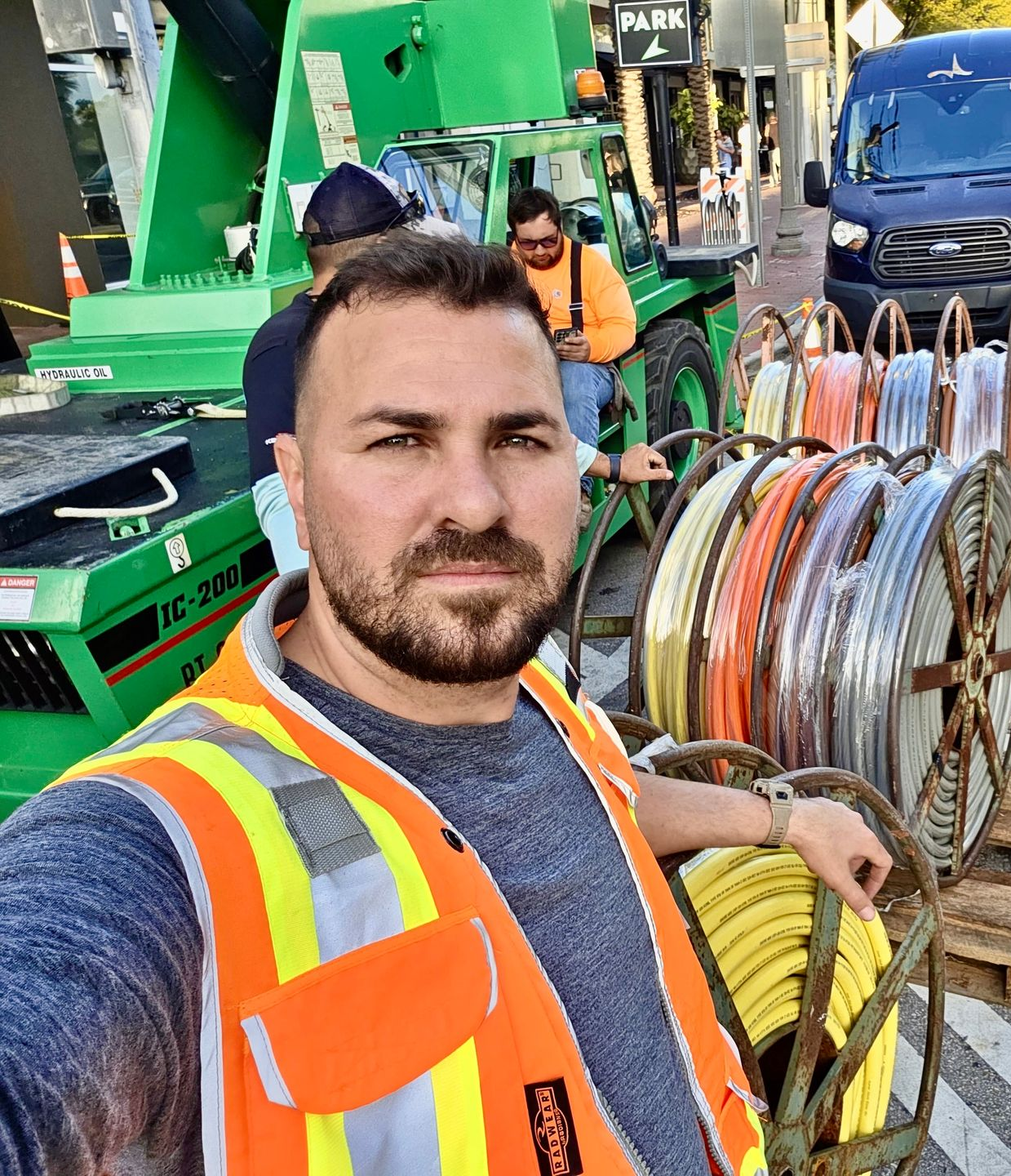 A person in a high-visibility orange safety vest poses for a selfie at a construction site with reels of wire and a crane.