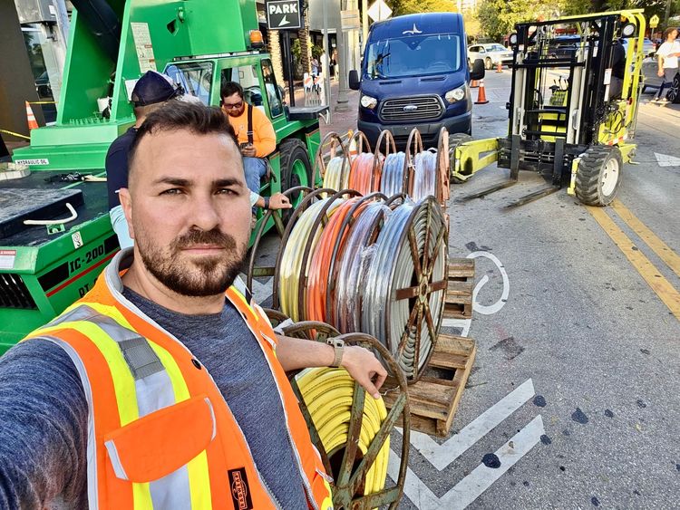 A construction worker wearing a high-visibility vest takes a selfie on a street with cable spools and heavy machinery.