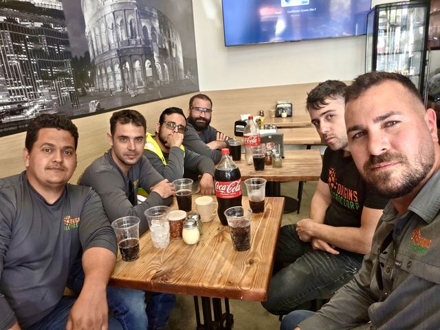 Six people sit together at a wooden restaurant table, posing for a selfie in front of a large wall mural.
