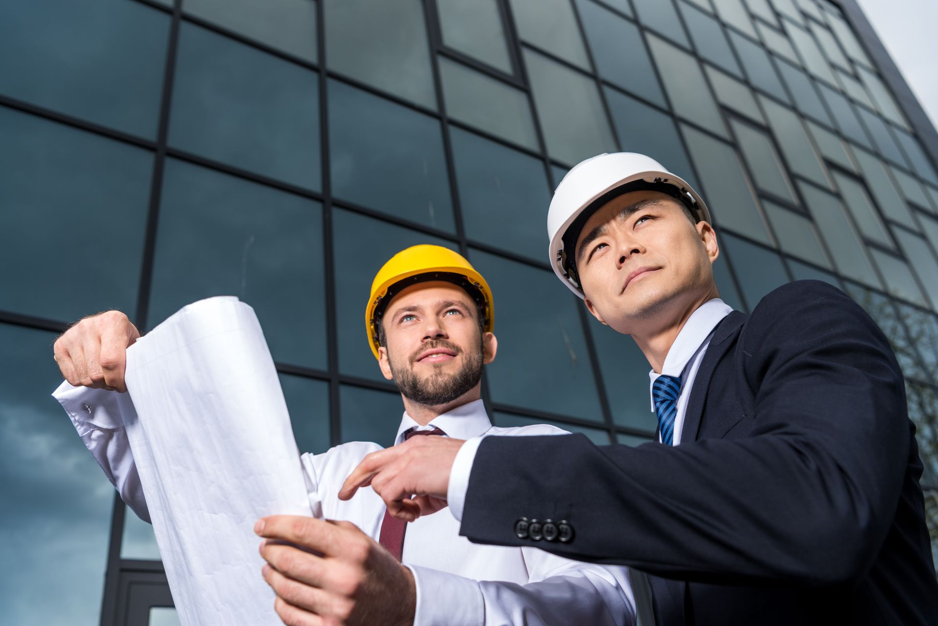 A construction worker in a white hard hat and green overalls stands with arms crossed against a plain grey background.