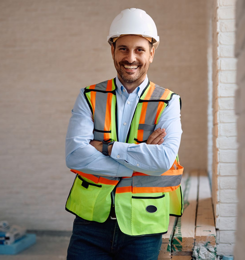 A construction worker in a white hard hat and green overalls stands with arms crossed against a plain grey background.