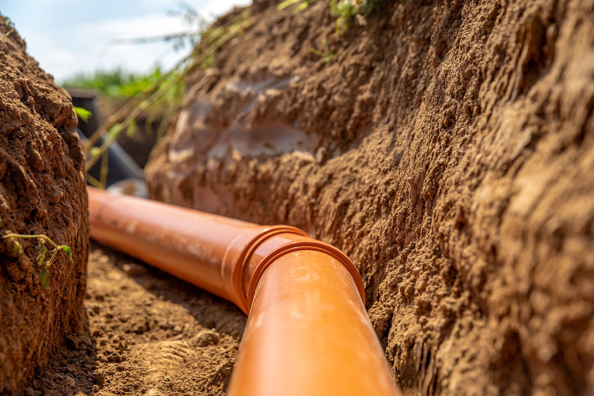 An orange PVC sewer pipe lies at the bottom of a narrow, freshly dug dirt trench.