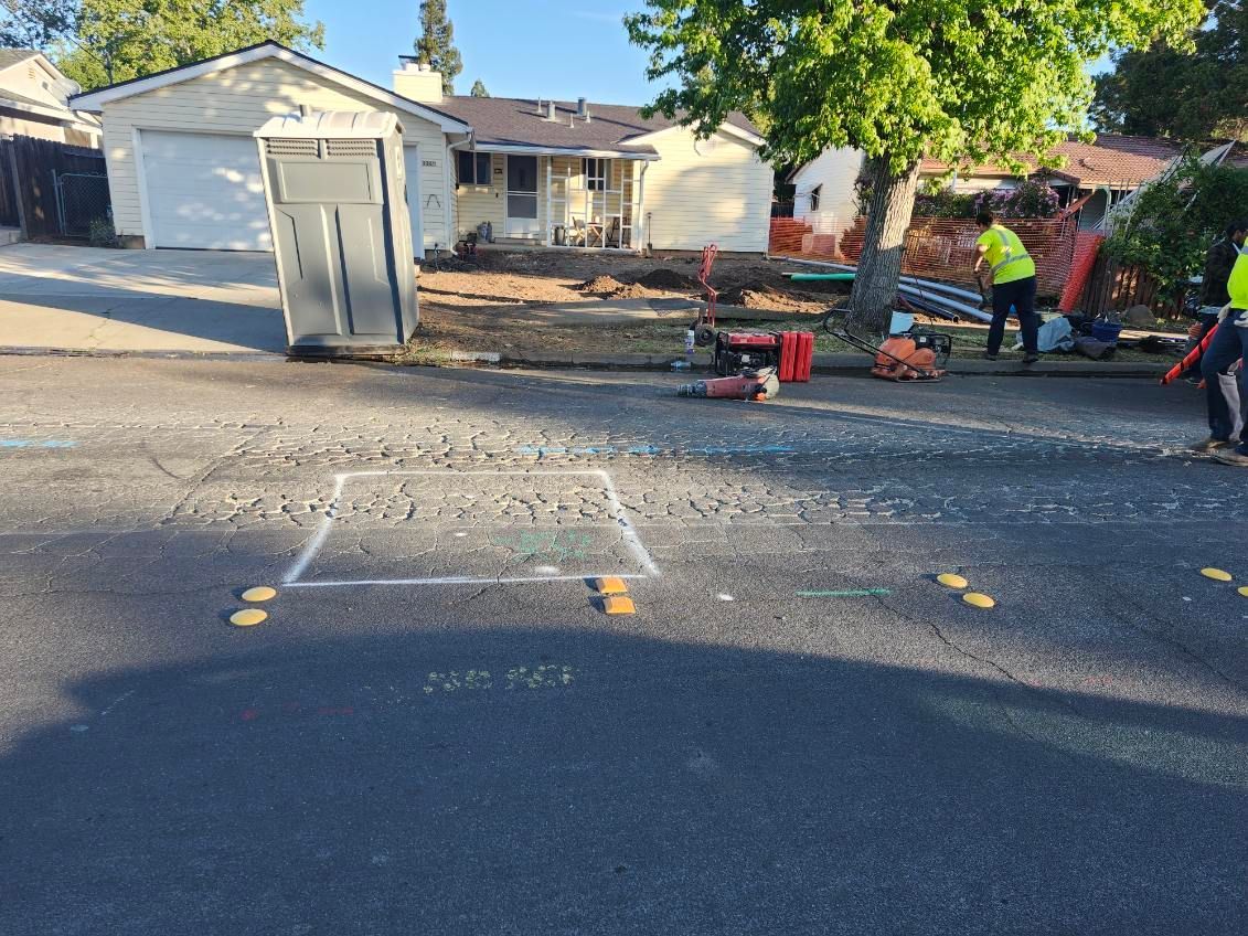 Street repair scene: workers in yellow vests, a white box outline on asphalt, and tools, with houses in the background.