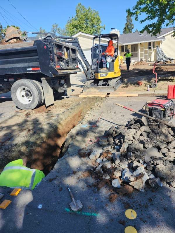 Construction site with a mini-excavator digging in a street. A dump truck is next to a deep trench in the pavement.