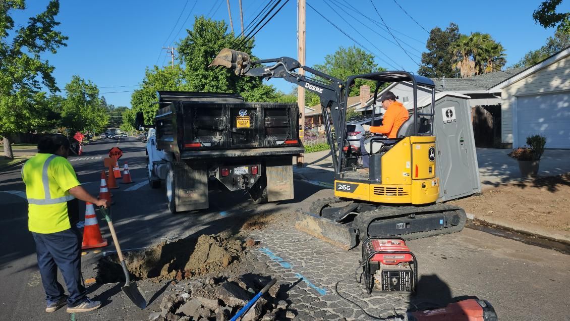 Road construction: Workers using a mini excavator and truck to dig on a street. One worker in a safety vest stands nearby.
